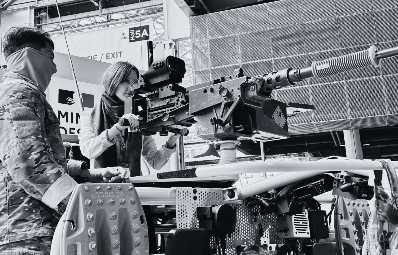 A soldier in camouflage and a colleague handling a vehicle-mounted weapon system at a defense exhibition.